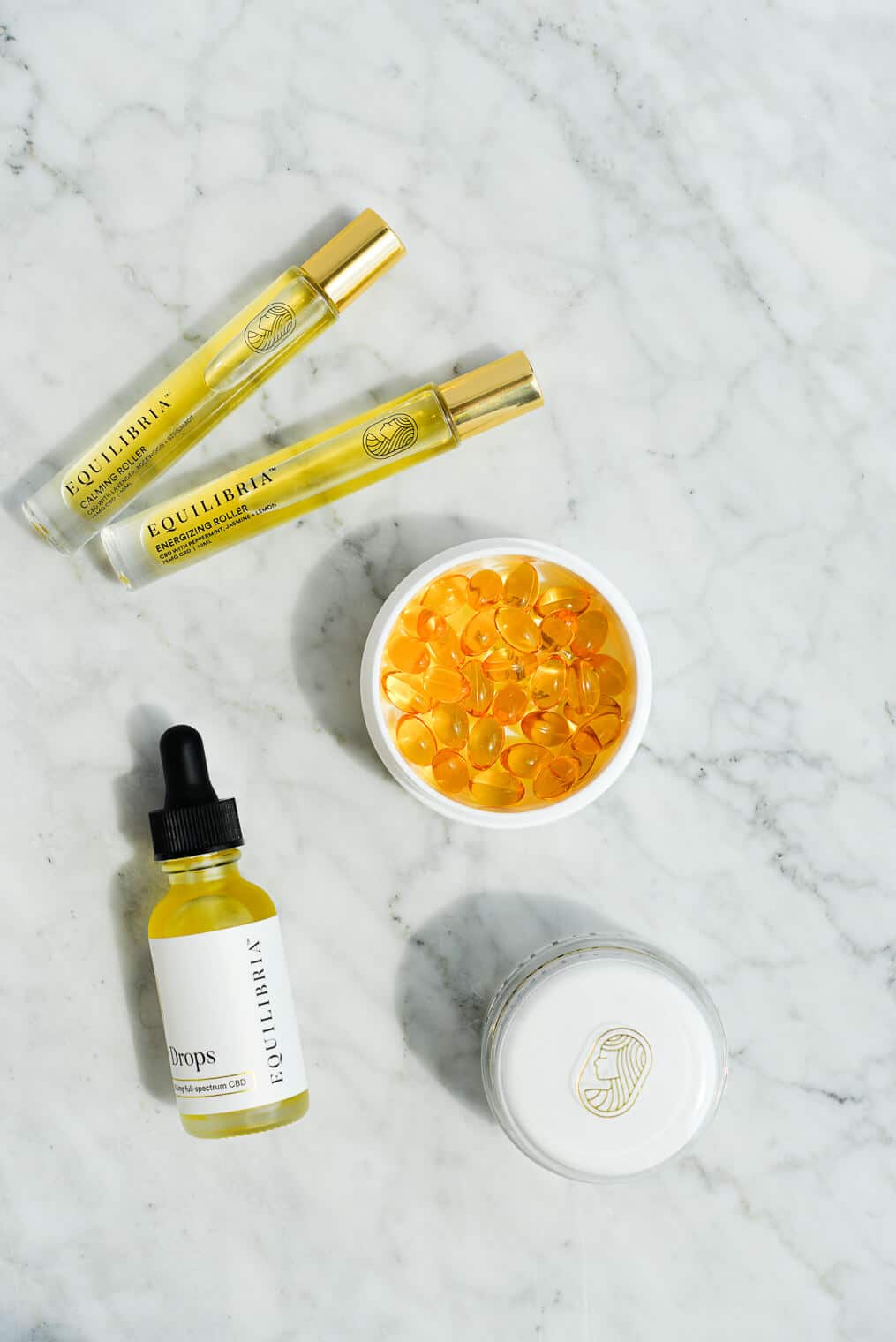 overhead shot of a glass bottle of equilibria cbd drops, rollers, relief cream, and softgels on a marble surface