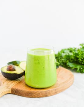 a small clear glass filled to the top with a low carb green smoothie sitting on a wood surface next to an avocado and a bunch of kale