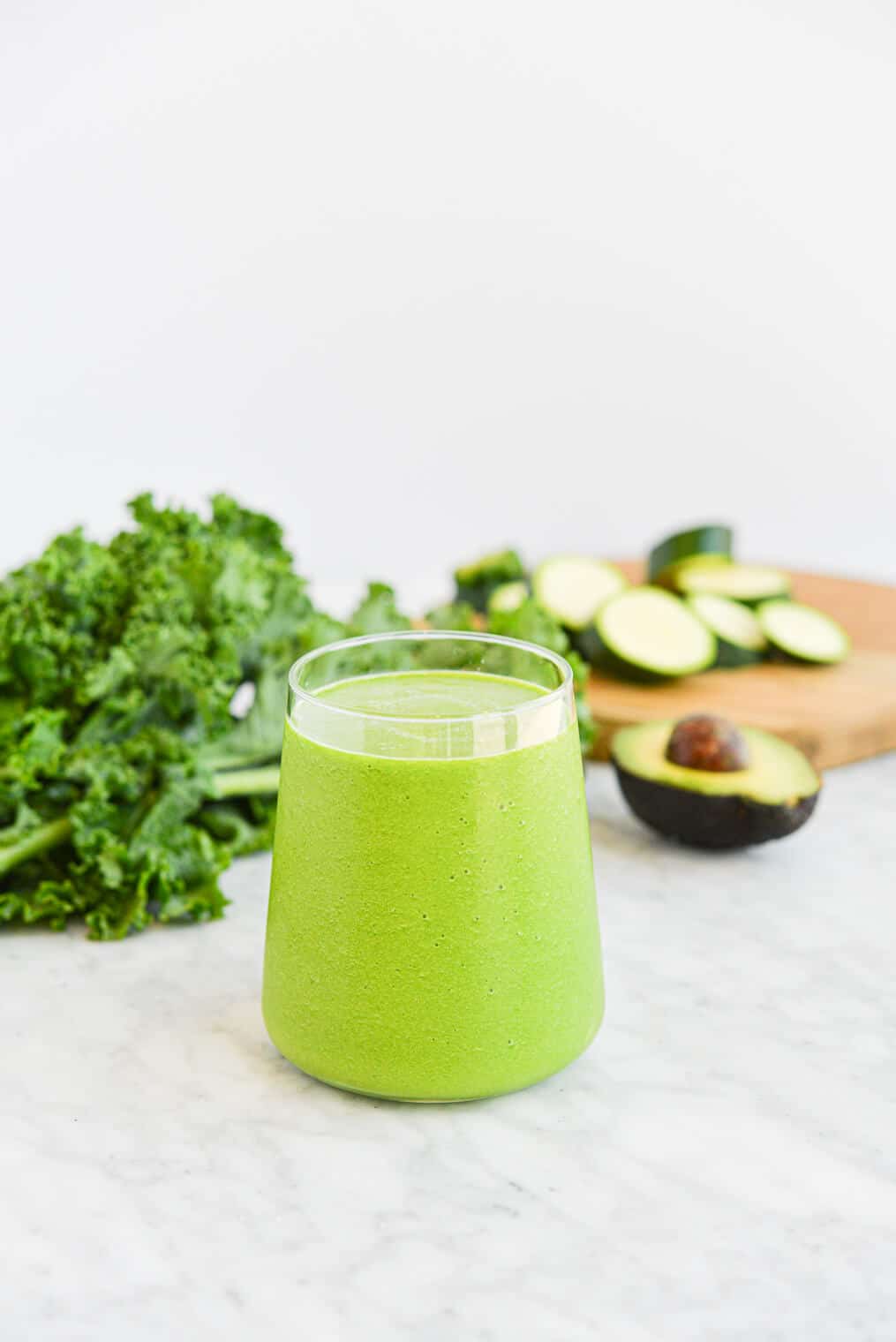 a small clear glass filled to the top with a low carb green smoothie sitting on a marble surface next to an avocado and a bunch of kale