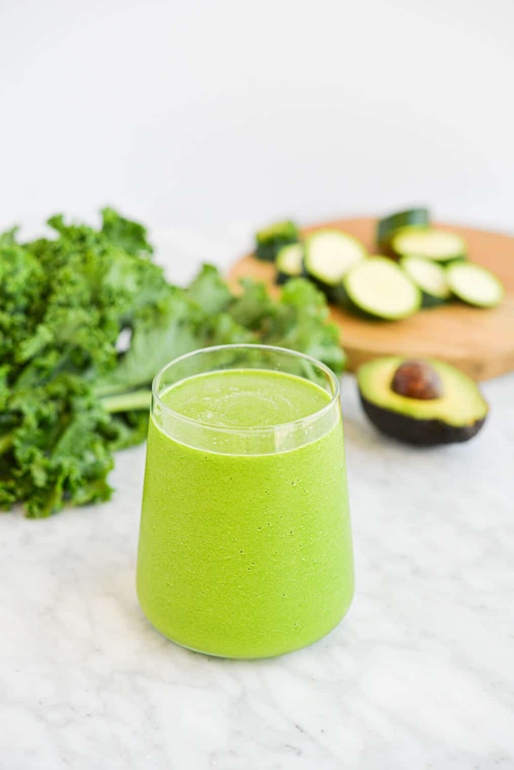 a small clear glass filled to the top with a low carb green smoothie sitting on a marble surface next to an avocado and a bunch of kale