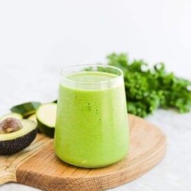 a small clear glass filled to the top with a low carb green smoothie sitting on a wood surface next to an avocado and a bunch of kale