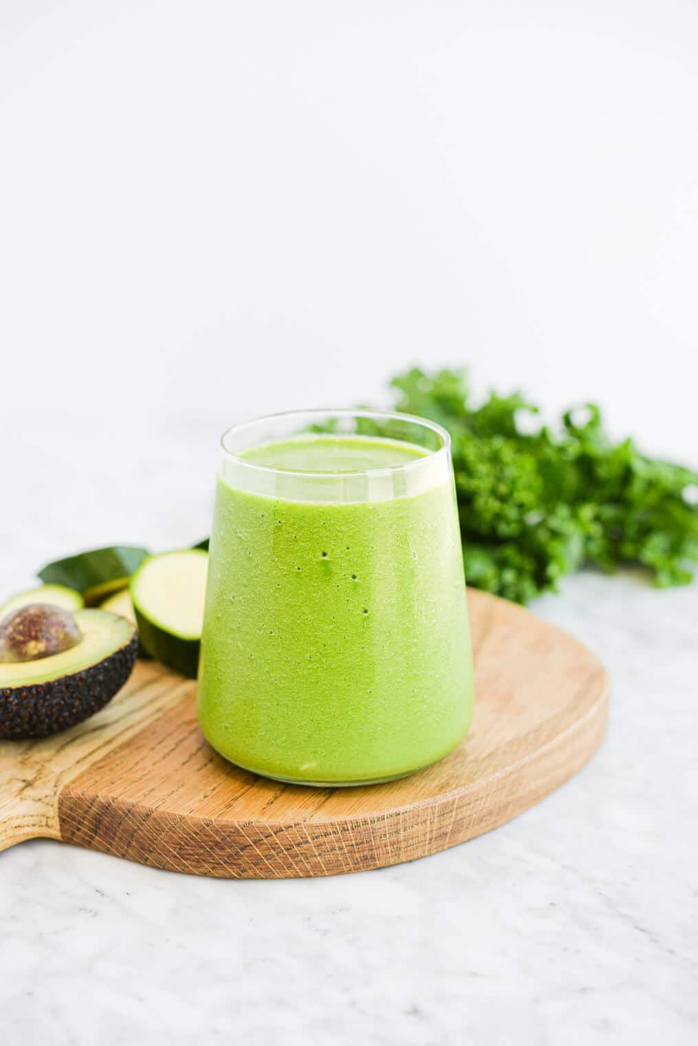 a small clear glass filled to the top with a low carb green smoothie sitting on a wood surface next to an avocado and a bunch of kale