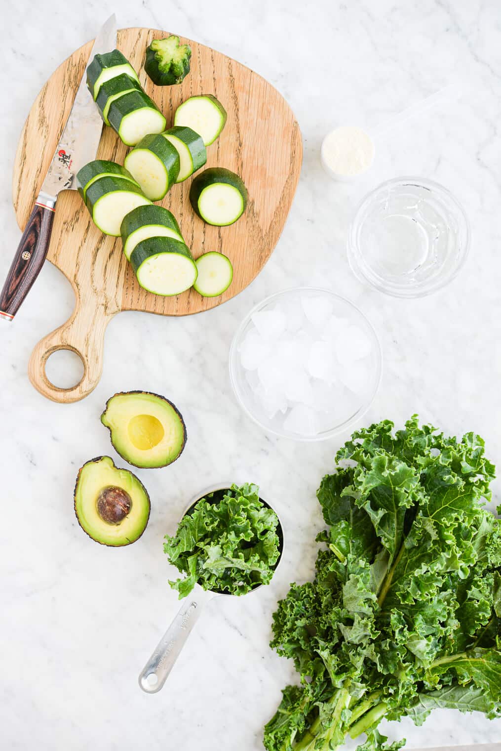 top view of the ingredients for low carb smoothies (kale, collagen peptides, zucchini, and avocado) sitting on a marble counter