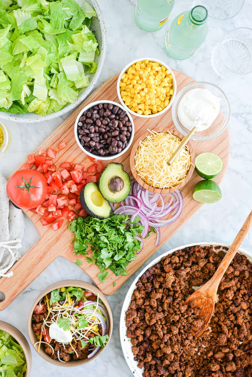 wooden cutting board on a marble surface topped with bowls of sour cream, shredded cheese, black beans, corn, diced tomatoes, cilantro, avocado, and red onion, next to a pan of ground beef