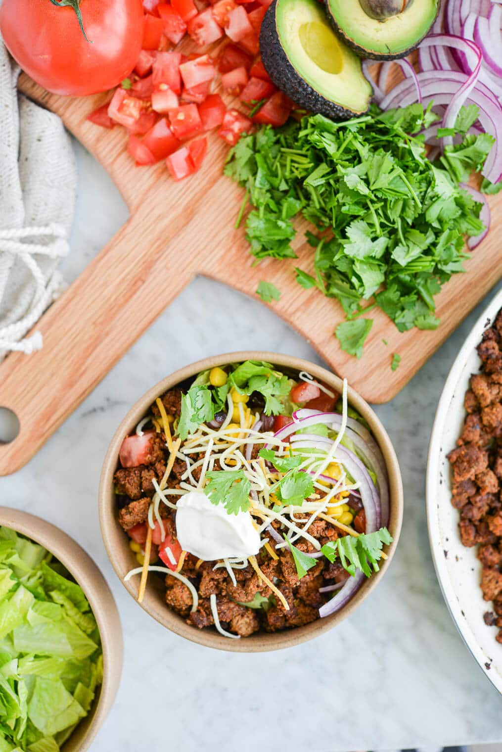 taco salad on a marble surface next to a wooden cutting board topped with chopped cilantro, a halved avocado, sliced red onion, and diced tomatoes