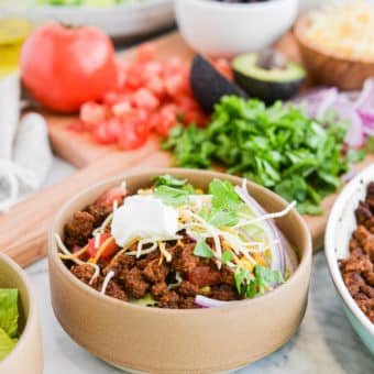 taco salad on a marble surface next to a wooden cutting board topped with chopped cilantro, a halved avocado, sliced red onion, and diced tomatoes