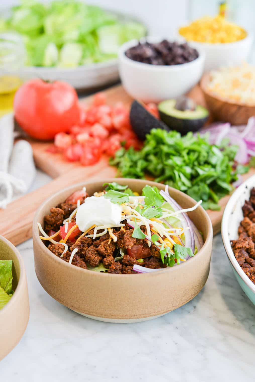 taco salad on a marble surface next to a wooden cutting board topped with chopped cilantro, a halved avocado, sliced red onion, and diced tomatoes