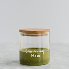 dark green matcha and clay peel off mask in a glass jar with a wooden lid with clarifying mask written on the jar in white lettering on a marble surface