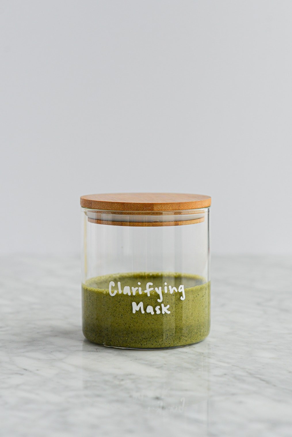 dark green matcha and clay peel off mask in a glass jar with a wooden lid with clarifying mask written on the jar in white lettering on a marble surface