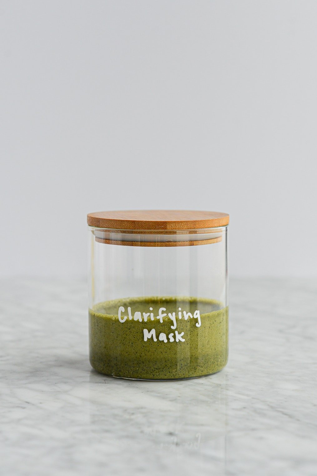 Mascarilla exfoliante de matcha y arcilla de color verde oscuro en un tarro de cristal con tapa de madera con mascarilla clarificante escrita en el tarro con letras blancas sobre una superficie de mármol