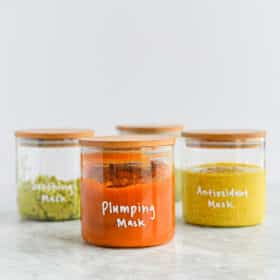 pumpkin face mask in a glass jar with a wooden lid with plumping mask written on the jar in white letters in front of 3 other glass jars full of masks on a marble surface