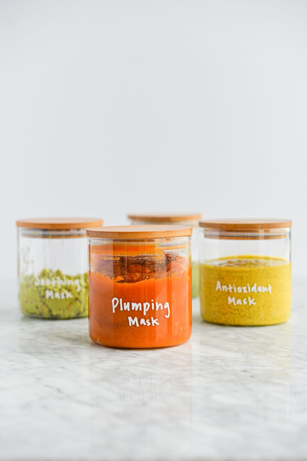 pumpkin face mask in a glass jar with a wooden lid with plumping mask written on the jar in white letters in front of 3 other glass jars full of masks on a marble surface