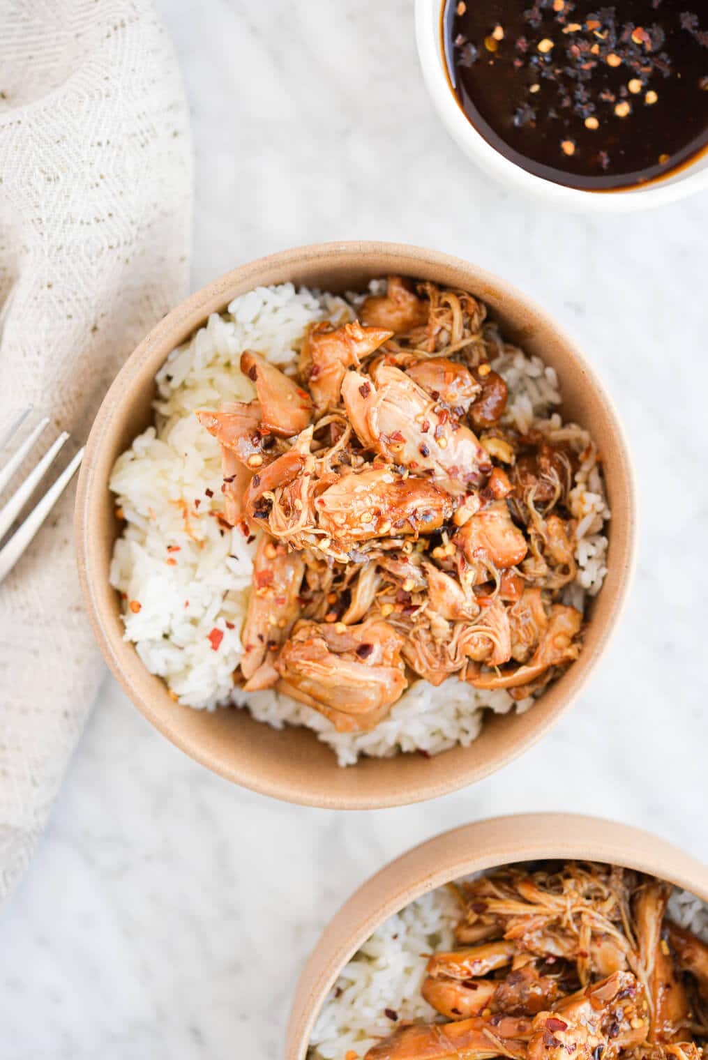 a beige bowl filled with white rice and shredded instant pot bourbon chicken with red pepper flakes on top on a marble surface with a linen napkin beside it