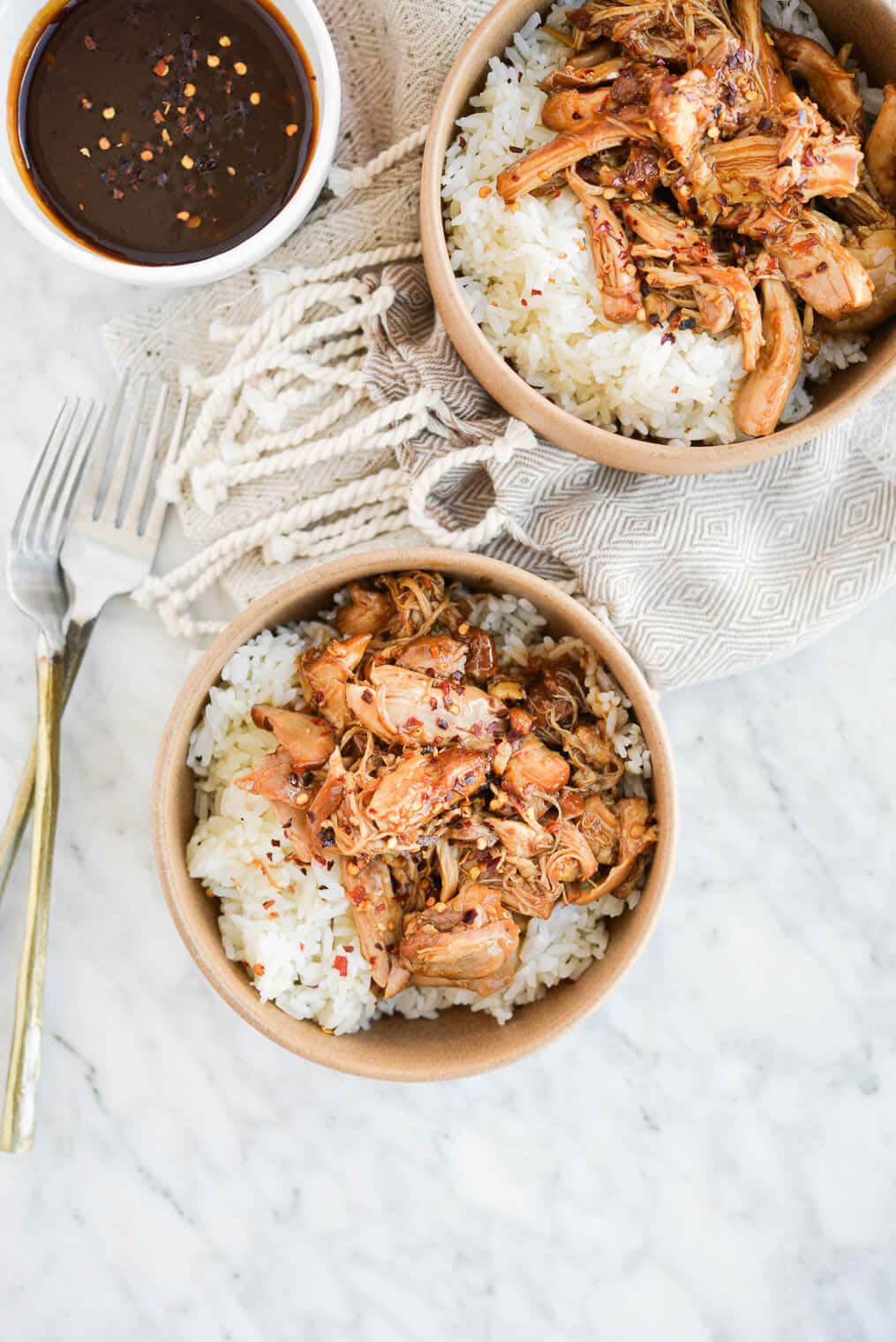 two beige bowls filled with white rice topped with shredded bourbon chicken with red pepper flakes on a marble surface