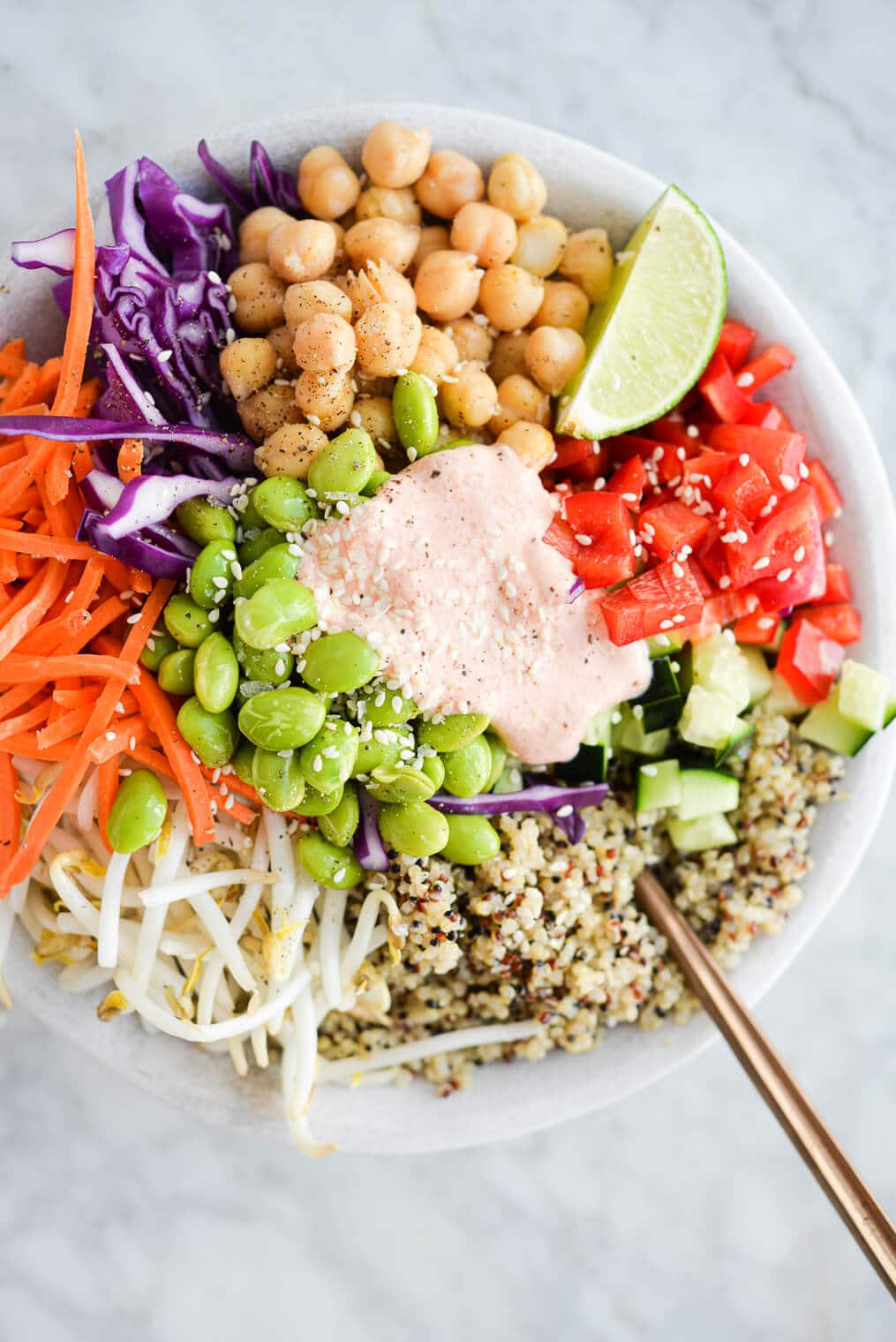 a close up of a thai red curry buddha bowl filled with quinoa, colorful, raw veggies, and a dollop of creamy yogurt red curry sauce on top, all sitting on a marble surface