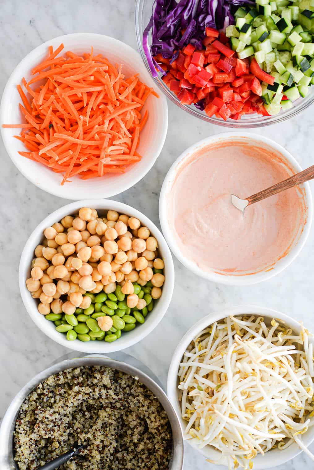 the makings of a thai red curry buddha bowl (a bowl of quinoa, mug bean sprouts, edamame and chickpeas, fresh, raw veggies, and a bowl of creamy thai red curry sauce sitting on a marble surface