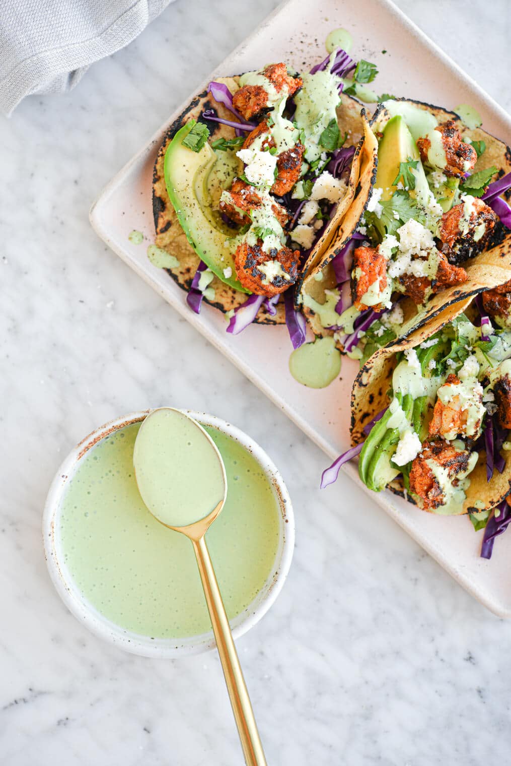 the top view of a platter of shrimp tacos (filled with seared shrimp, sliced avocado, red cabbage, and a creamy green yogurt sauce) sitting next to a bowl filled with more sauce all sitting on a marble surface