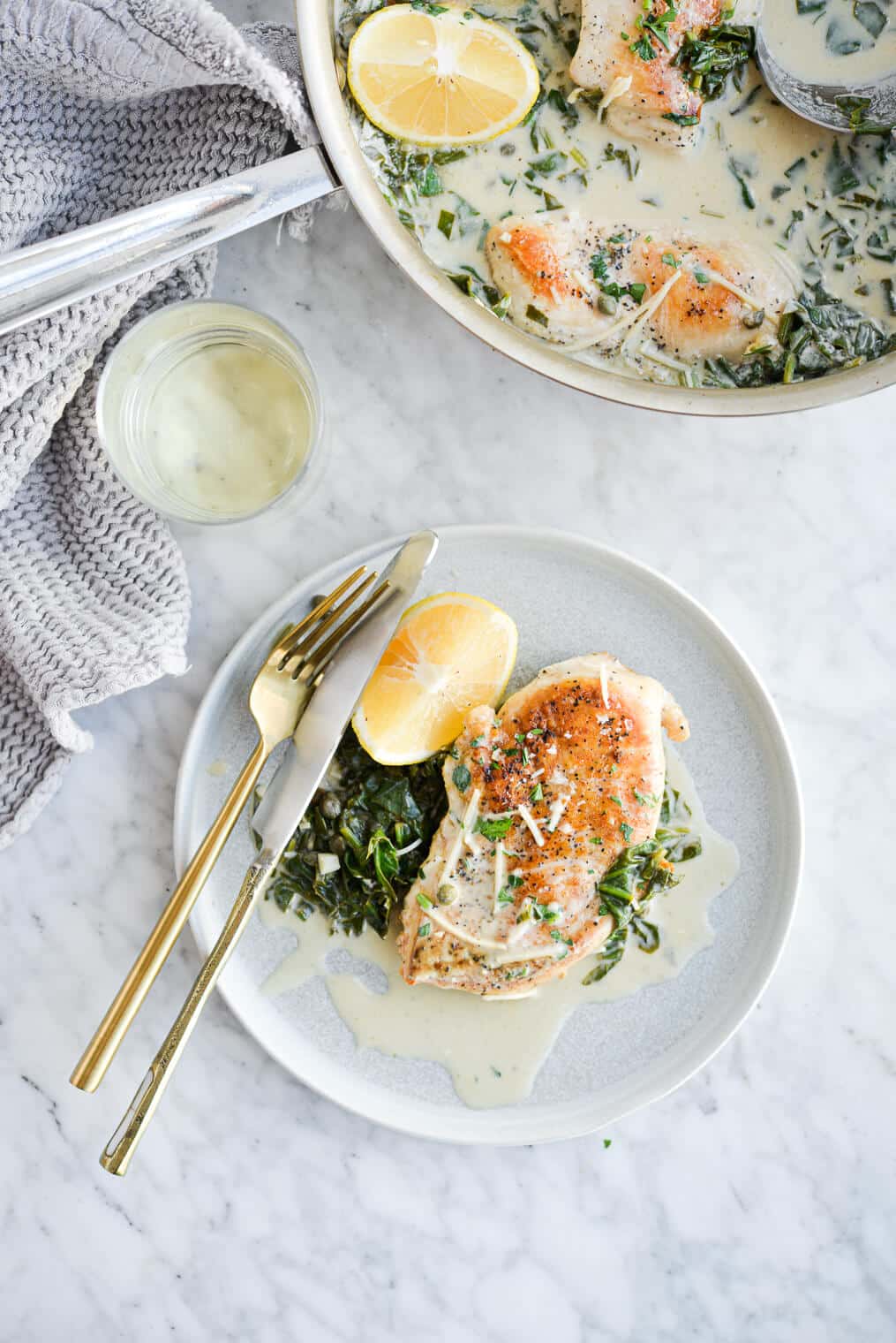 The top view of a plate of creamy chicken piccata sitting next to a glass of white wine and a skillet of chicken piccata all on a marble surface.