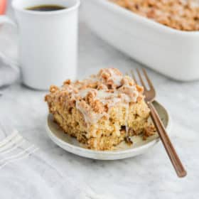 the side view of a piece of apple coffee cake on a plate sitting next to the dish of coffee cake, a mug of black coffee, and a basket of apples