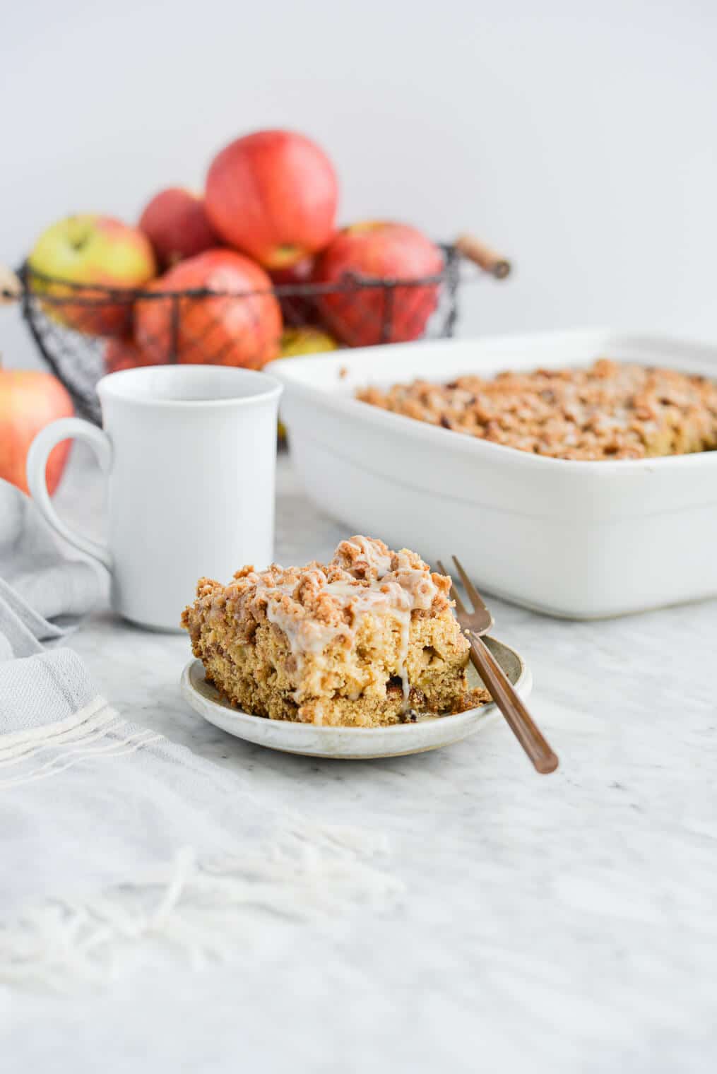 the side view of a piece of apple coffee cake on a plate sitting next to the dish of coffee cake, a mug of black coffee, and a basket of apples