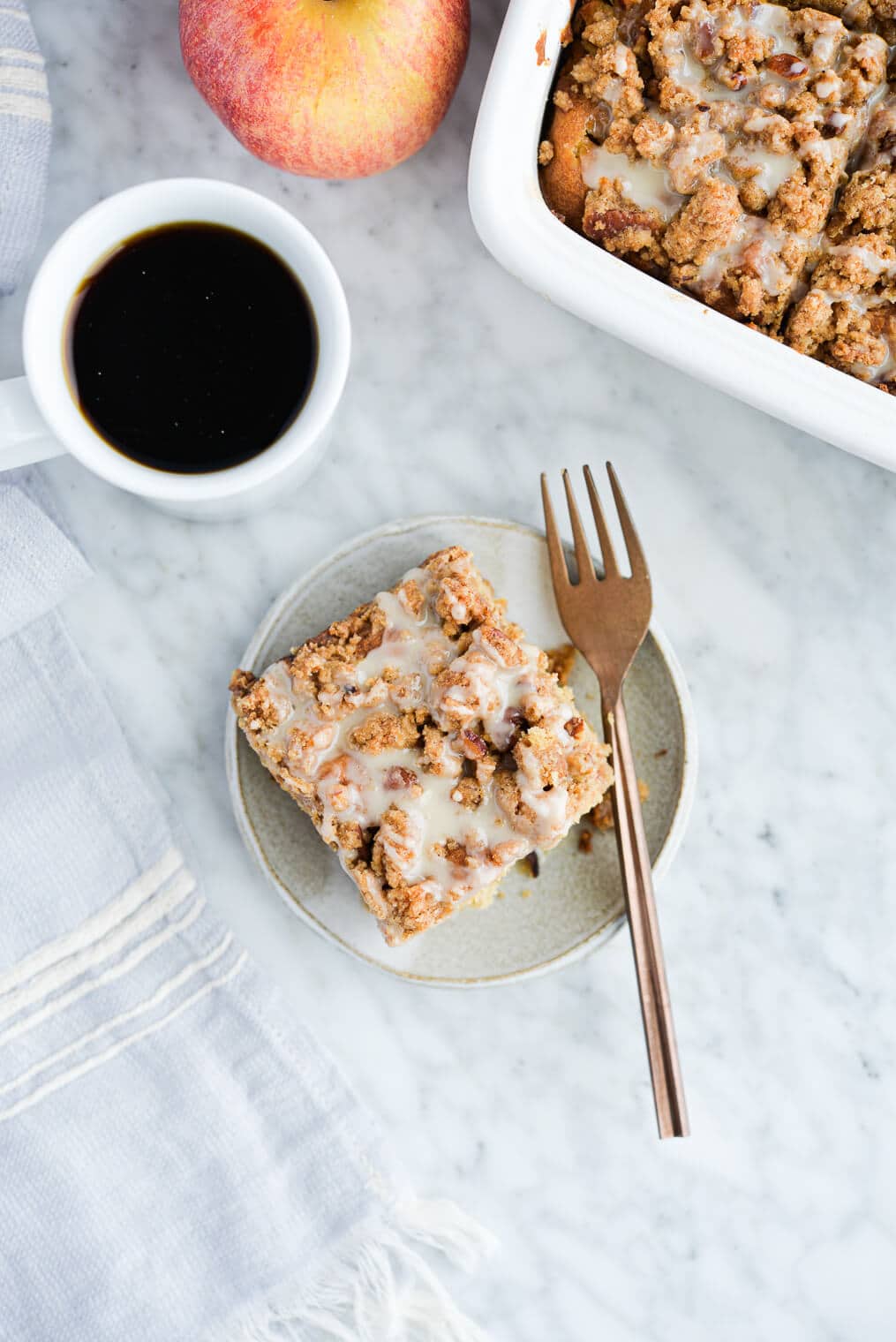 the top view of a piece of apple coffee cake on a plate sitting next to the dish of coffee cake and a mug of black coffee