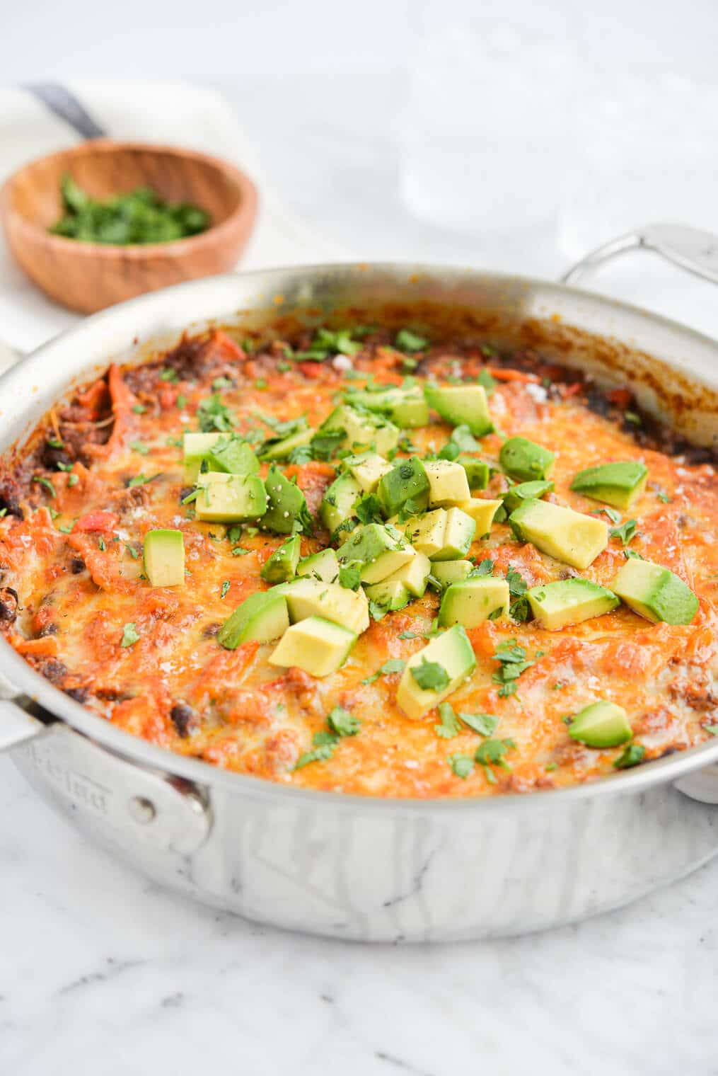 the side view of a skillet of ground beef enchilada casserole topped with diced avocado and cilantro sitting on a marble surface