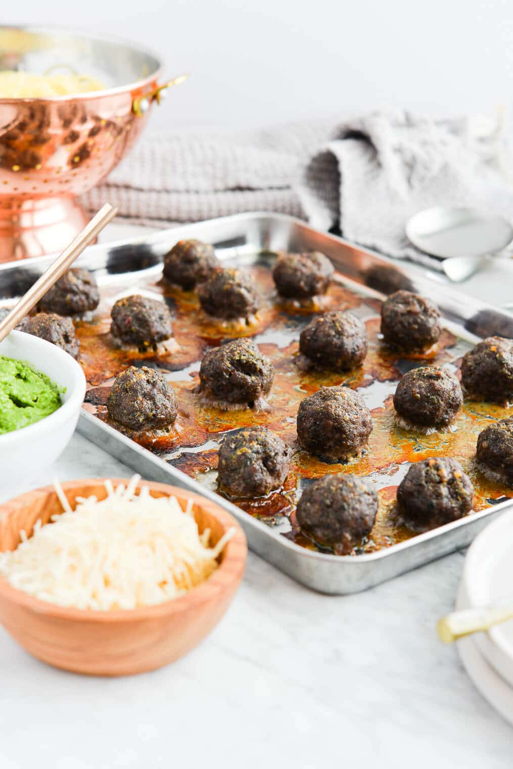 the side view of a sheet pan lined with cooked pesto meatballs sitting next to a bowl of green pesto, a small bowl of parmesan cheese, and a bowl of cooked noodles all on a marble surface