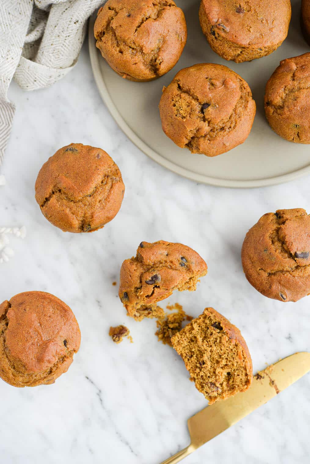 the top view of a plate of pumpkin chocolate chip muffins sitting on a marble countertop next to 4 pumpkin muffins, one of which has been sliced open with a gold butter knife
