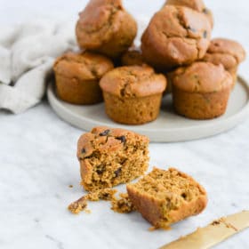 the side view of a batch of pumpkin chocolate chip muffins sitting on a plate next to one muffin on the countertop that has been sliced in half with a gold butter knife