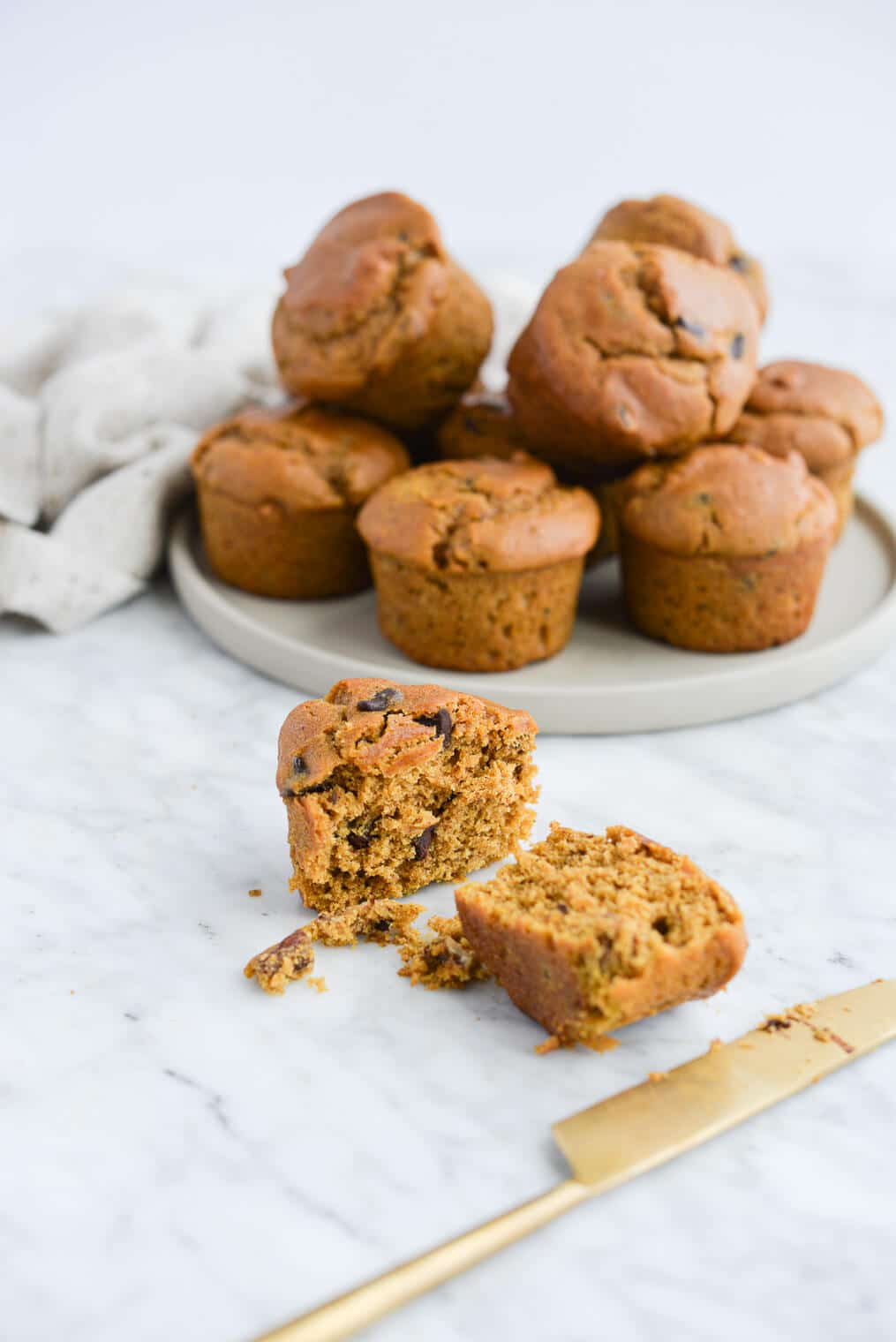the side view of a batch of pumpkin chocolate chip muffins sitting on a plate next to one muffin on the countertop that has been sliced in half with a gold butter knife