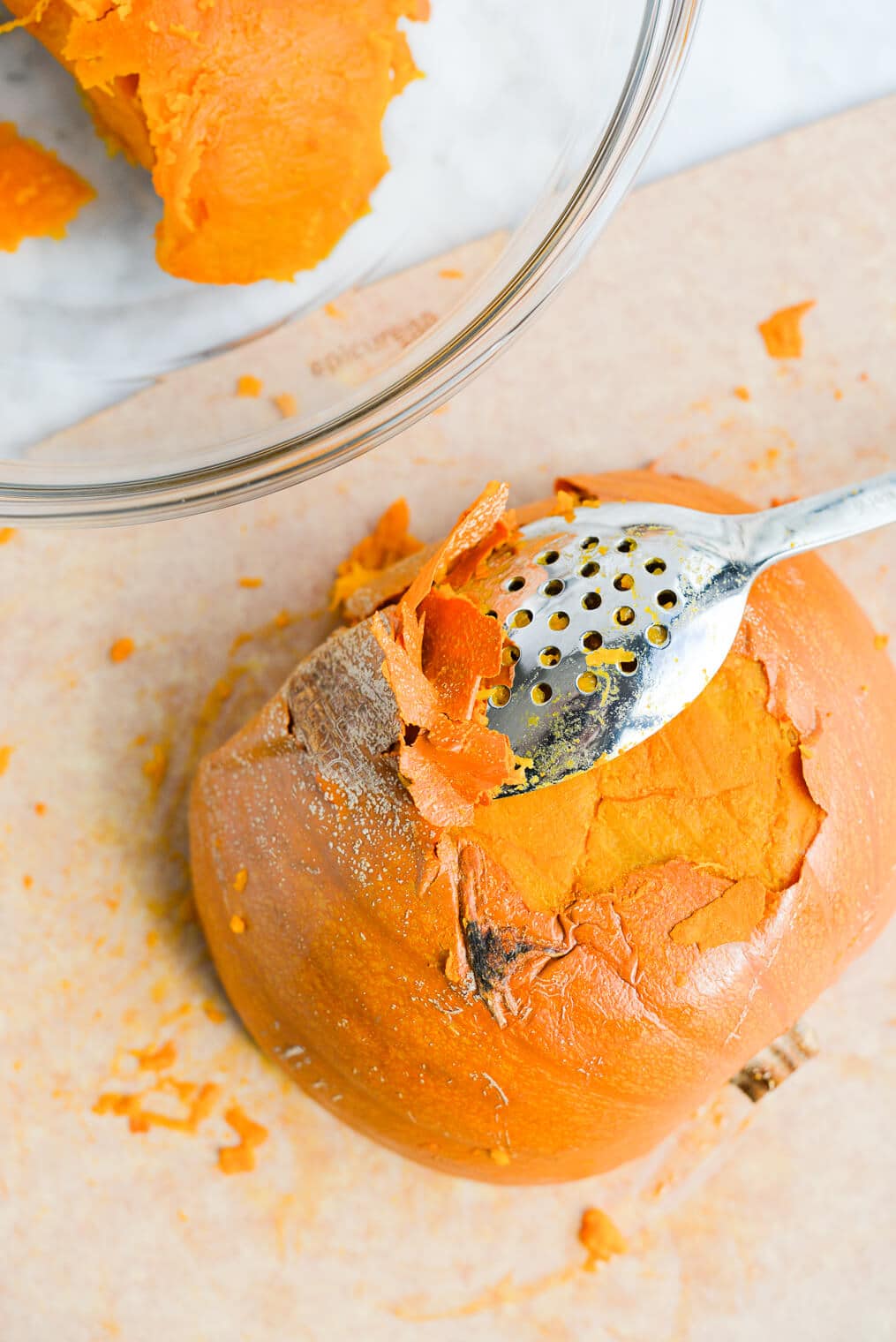 a halved cooked pumpkin with a blunt-nose spoon peeling off part of the pumpkin skin and a bowl of scooped out pumpkin flesh