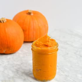 homemade pumpkin puree filling a mason jar sitting in front of two baking pumpkins on a marble surface