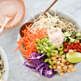 a close up of a thai red curry buddha bowl filled with quinoa, colorful, raw veggies, and a dollop of creamy yogurt red curry sauce on top, all sitting on a marble surface