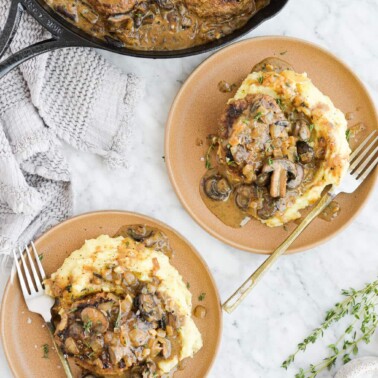 the top view of two plates of salisbury steak, mashed potatoes, and mushroom gravy sitting next to a skillet of salisbury steak on a marble surface