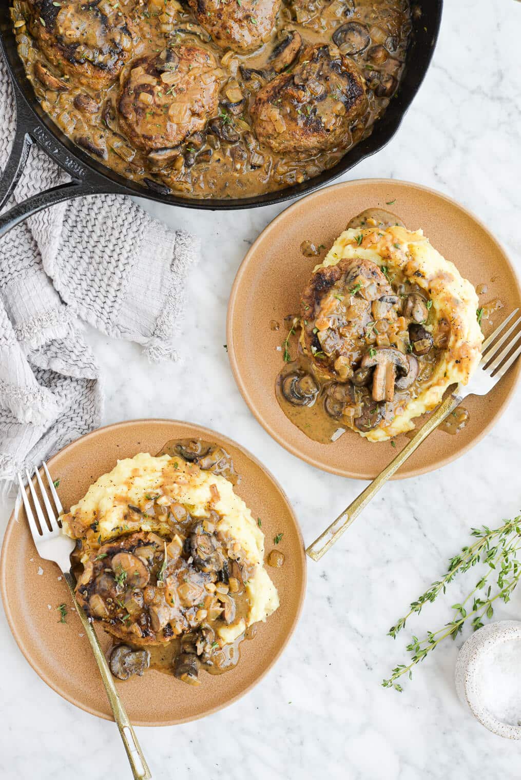 the top view of two plates of salisbury steak, mashed potatoes, and mushroom gravy sitting next to a skillet of salisbury steak on a marble surface