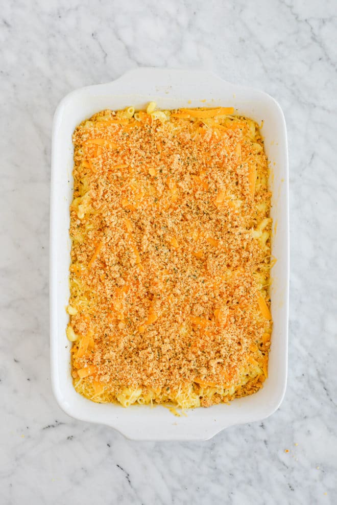 the top view of a casserole dish of cooked macaroni noodles, shredded cheese, and topped with breadcrumbs, all sitting on a marble surface