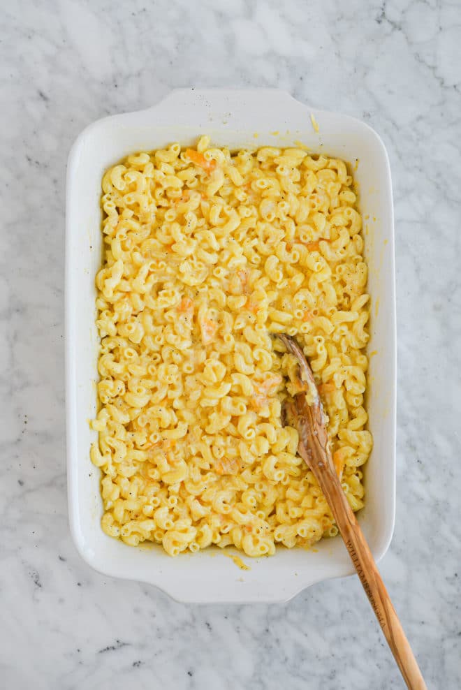 the top view of a casserole dish with cooked macaroni noodles and shredded cheese being stirred in with a wooden spoon all sitting on a marble surface