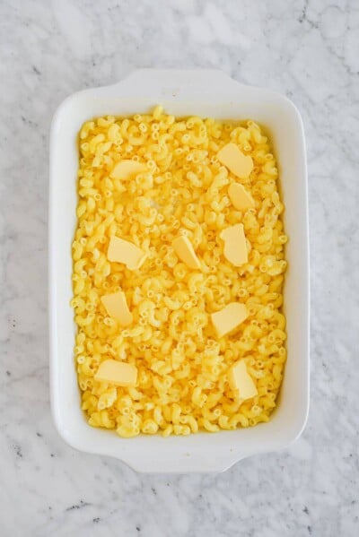 the top view of a casserole dish wish raw macaroni noodles and sliced butter sitting on a marble surface