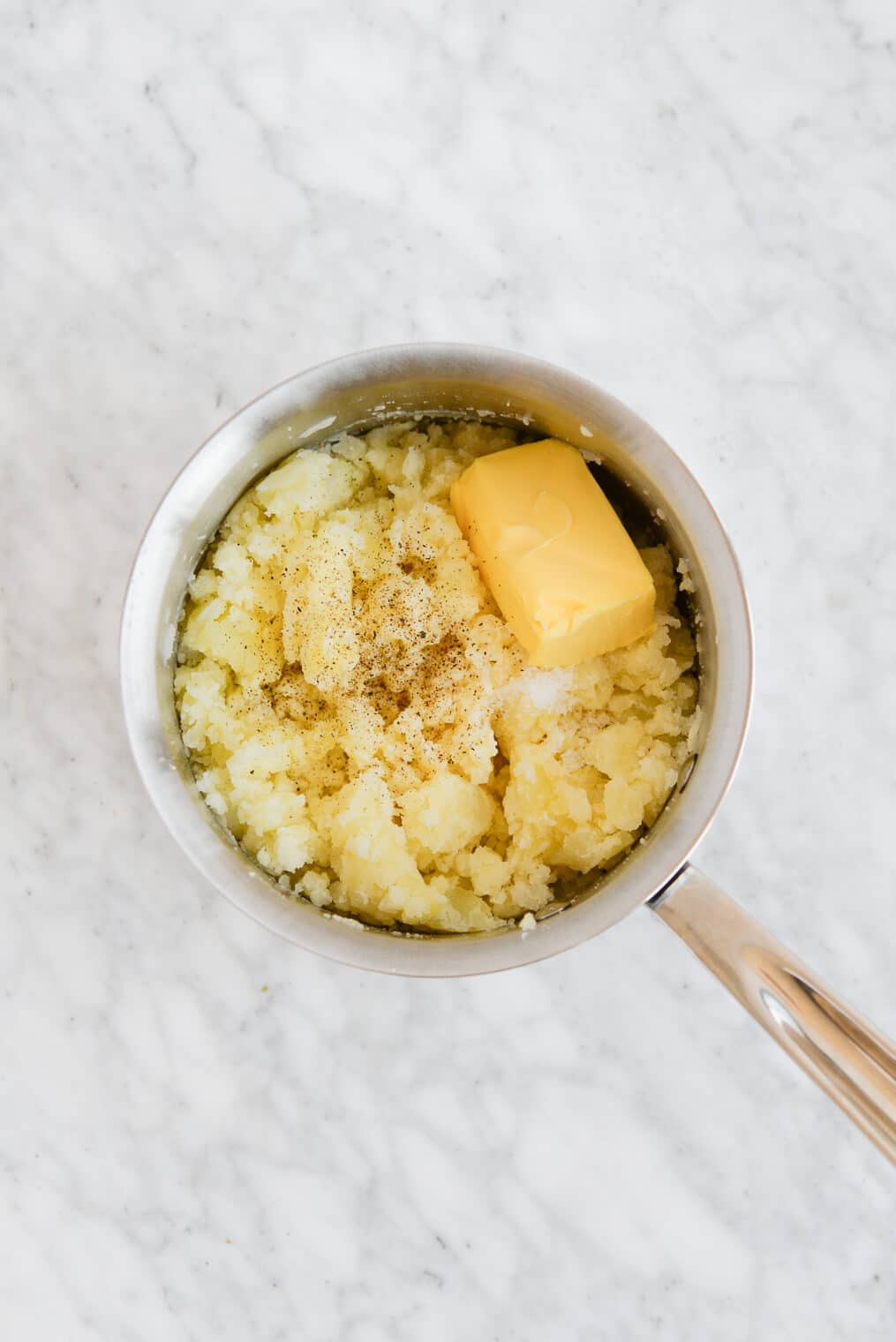 top view of a pot of mashed potatoes with a stick of butter melting on top of the mashed potatoes sitting on a marble surface