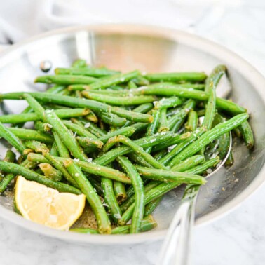 bright green sauteed green beans in a stainless steel pan next to a lemon wedge