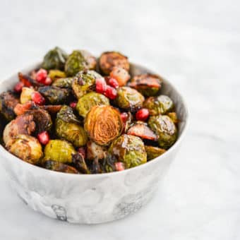 a bowl of roasted brussels sprouts, pomegranate seeds, and bacon in a marble bowl on a marble surface