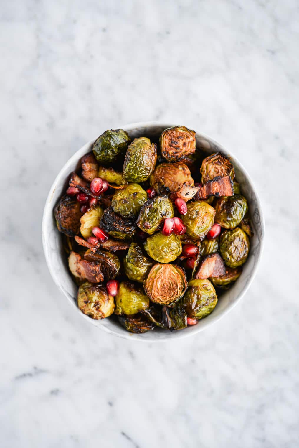 top view of a bowl of roasted brussels sprouts, pomegranate seeds, and bacon in a marble bowl on a marble surface