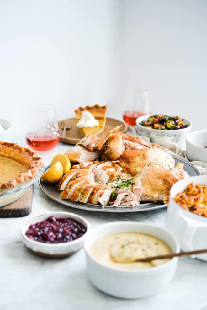 side view of a thanksgiving holiday spread complete with pumpkin pie, carved whole turkey, classic gravy, wine, cranberry sauce, brussels sprouts, and semi-homemade stuffing