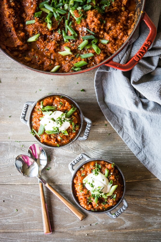 a large red dutch oven filled with a deep orange pumpkin chili topped with sliced green onions sitting next to two individual portions of chili topped with a dollop of sour cream and sliced green onions