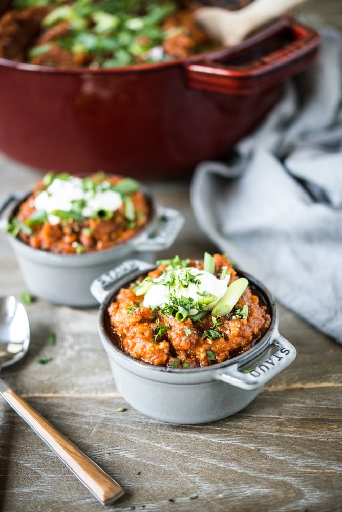 a large red dutch oven filled with a deep orange pumpkin chili topped with sliced green onions sitting next to two individual portions of chili topped with a dollop of sour cream and sliced green onions