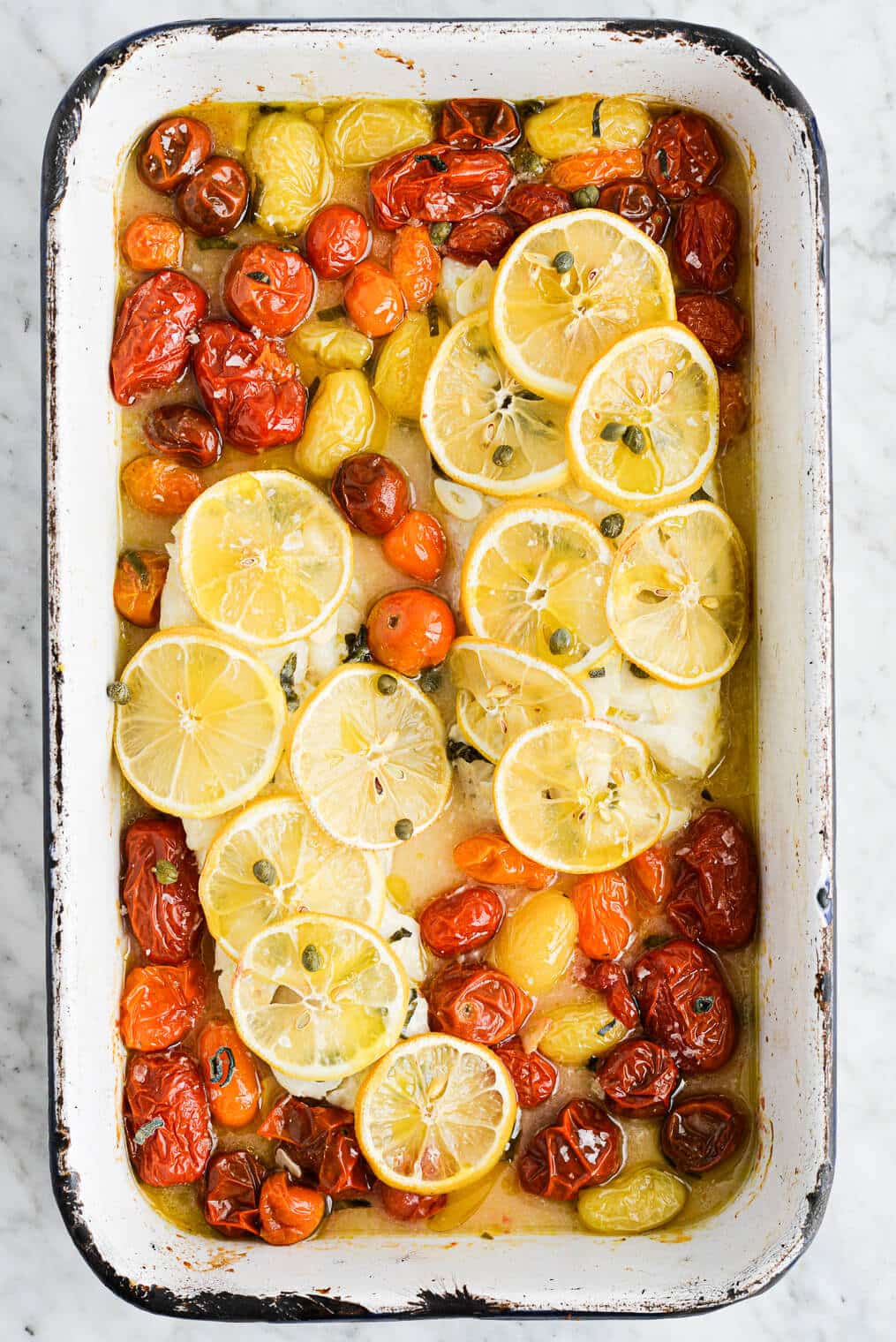 the top view of a roasting dish of baked cod topped with thin sliced lemons and roasted tomatoes