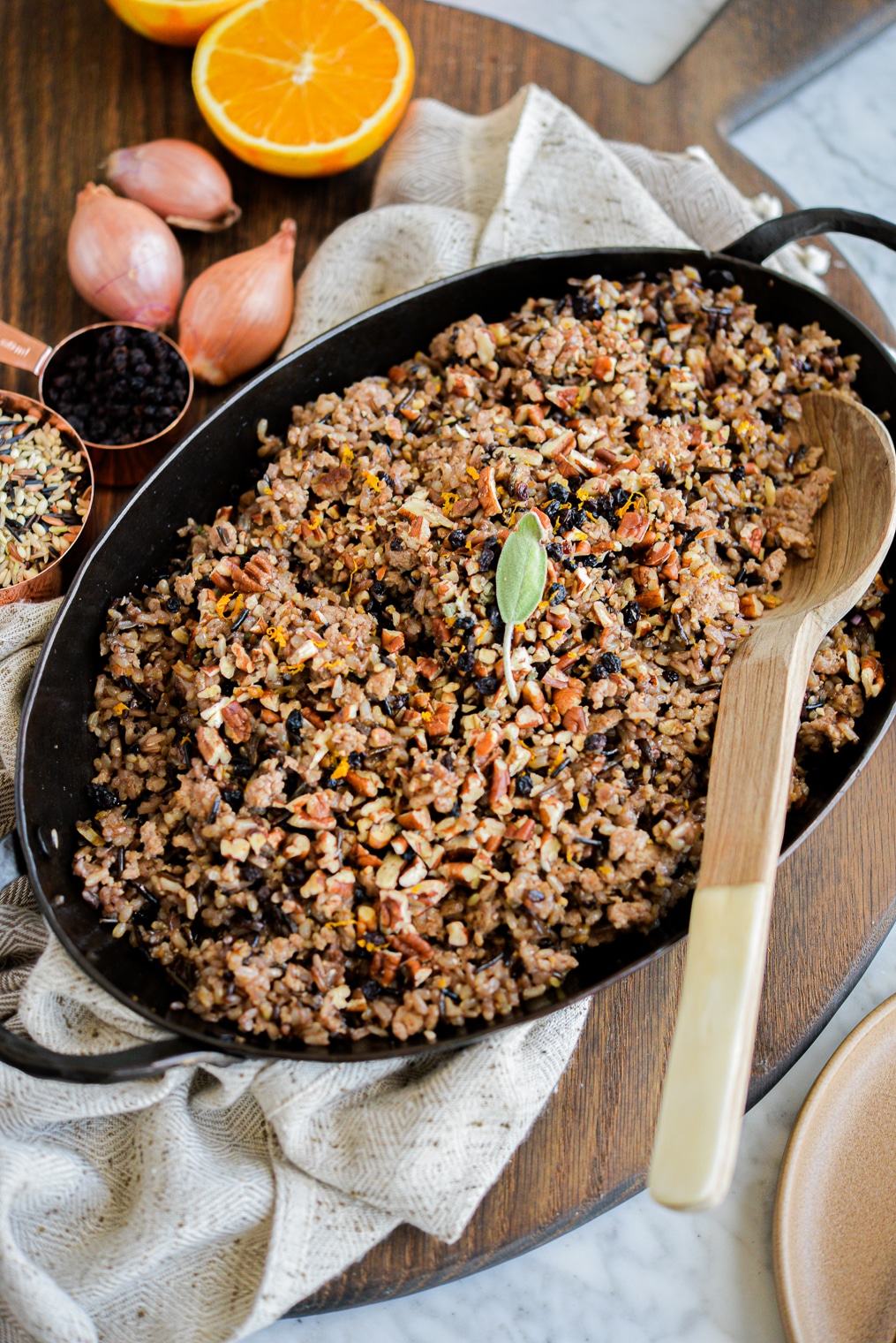 a dish of wild rice stuffing sitting on a wood board next to a halved orange, and whole shallots