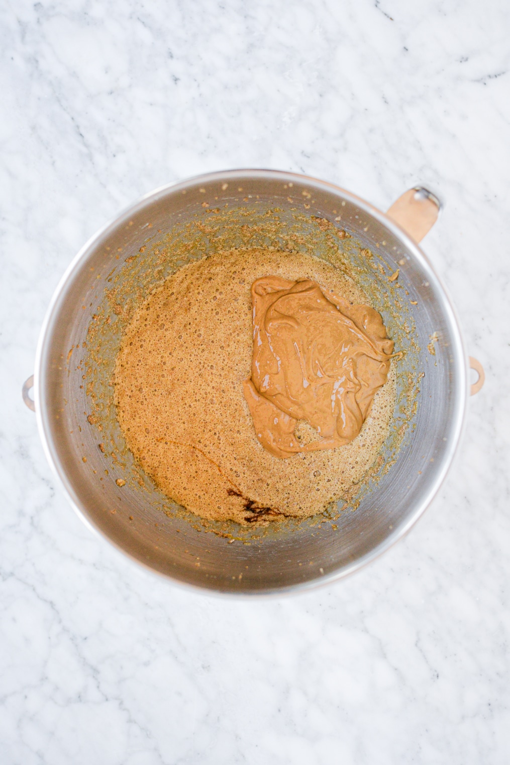 cashew butter being mixed into wet ingredients for ginger molasses cookies in a stainless steel bowl on a marble surface
