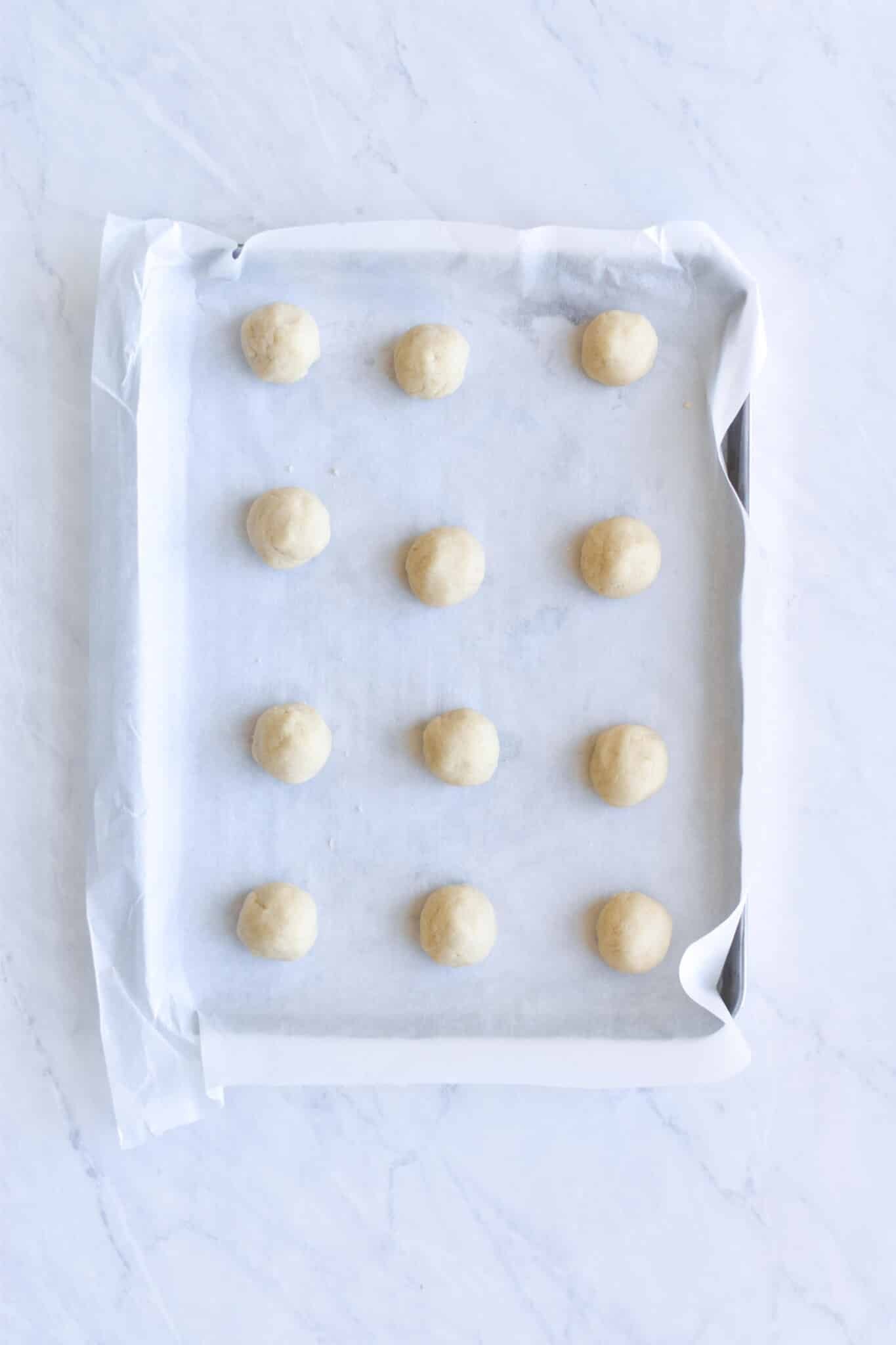 12 cookie dough balls on a parchment paper lined baking sheet on a marble surface