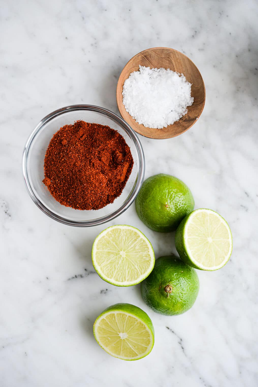 a small bowl of chili powder, a small bowl of sea salt, and two cut limes sitting on a marble surface